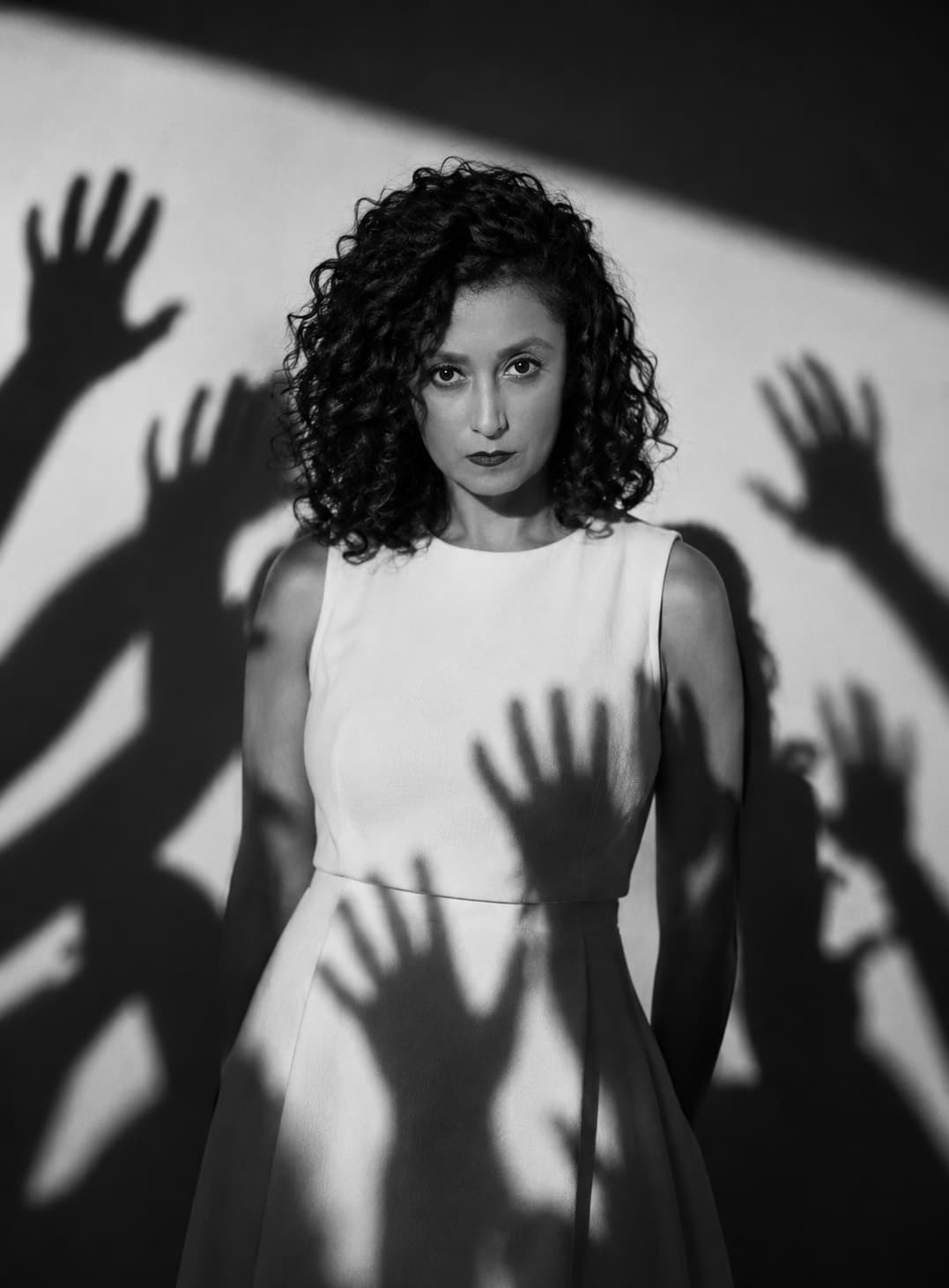 Florencia de vestido blanco frente a una pared cubierta de sombras de manos que se extienden hacia ella.